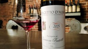 Wine glass of red wine beside a Dolcetto d'Alba 2025 bottle on a bar with a brick wall backdrop.