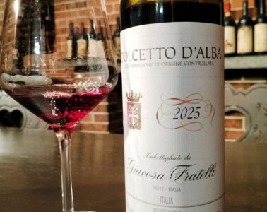 Wine glass of red wine beside a Dolcetto d'Alba 2025 bottle on a bar with a brick wall backdrop.