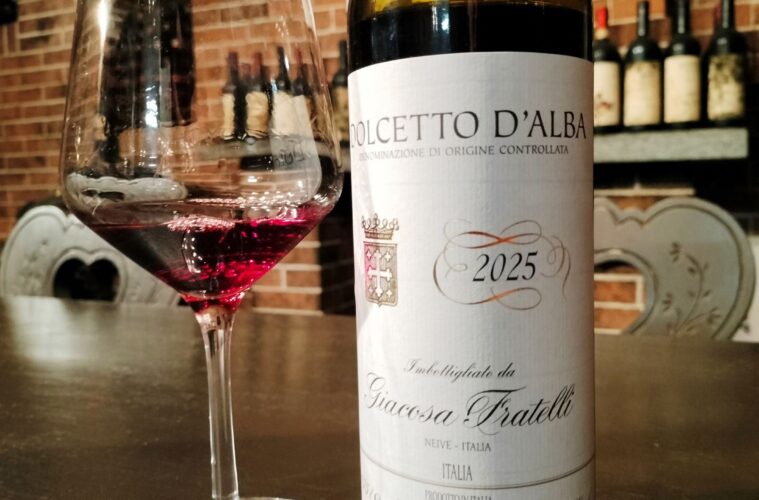 Wine glass of red wine beside a Dolcetto d'Alba 2025 bottle on a bar with a brick wall backdrop.