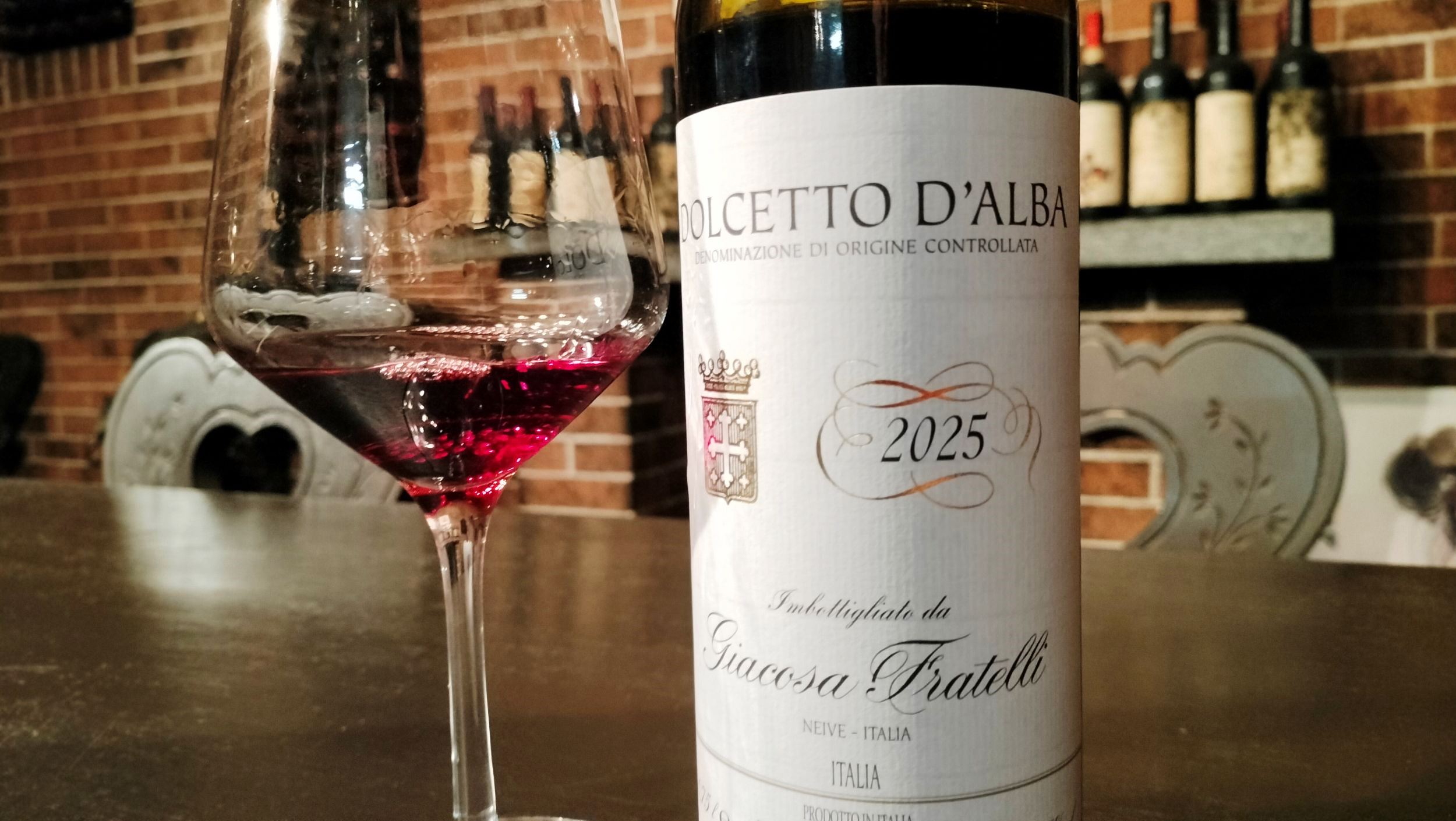 Wine glass of red wine beside a Dolcetto d'Alba 2025 bottle on a bar with a brick wall backdrop.