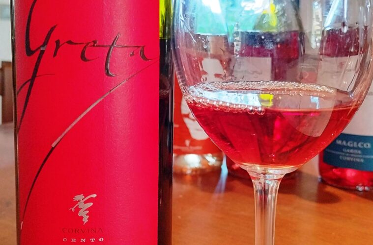 Close-up of a bright pink wine bottle label with cursive writing next to a glass of red wine. Note: label reads Corvina Cento Cento.
