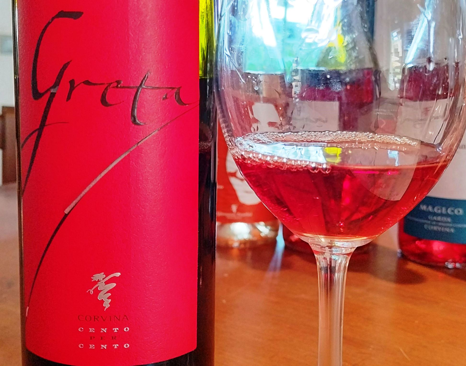 Close-up of a bright pink wine bottle label with cursive writing next to a glass of red wine. Note: label reads Corvina Cento Cento.