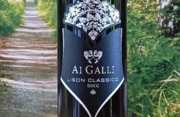 Bottle of AI GALLI Lison Classico DOCG wine with a black label and white crest against a garden backdrop; ornate crown at the top and a shield-shaped label showing the text AI GALLI LISON CLASSICO DOCG.