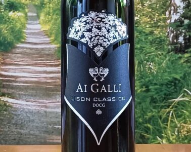 Bottle of AI GALLI Lison Classico DOCG wine with a black label and white crest against a garden backdrop; ornate crown at the top and a shield-shaped label showing the text AI GALLI LISON CLASSICO DOCG.