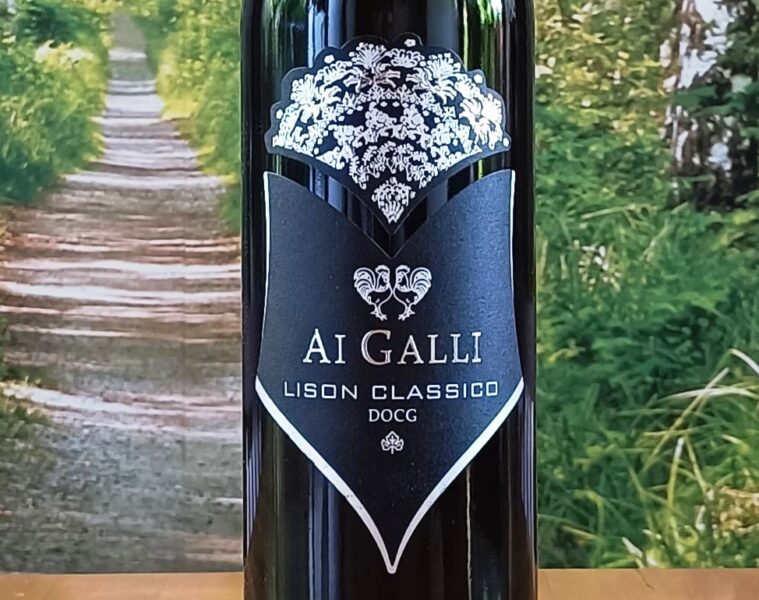 Bottle of AI GALLI Lison Classico DOCG wine with a black label and white crest against a garden backdrop; ornate crown at the top and a shield-shaped label showing the text AI GALLI LISON CLASSICO DOCG.
