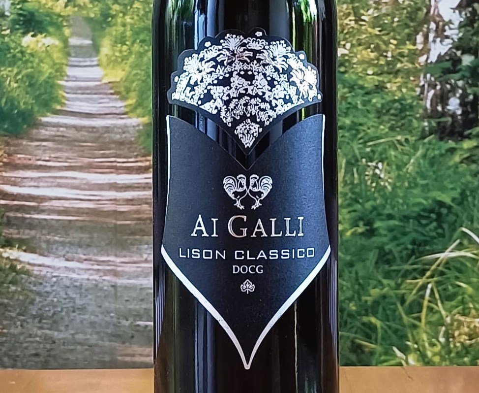 Bottle of AI GALLI Lison Classico DOCG wine with a black label and white crest against a garden backdrop; ornate crown at the top and a shield-shaped label showing the text AI GALLI LISON CLASSICO DOCG.