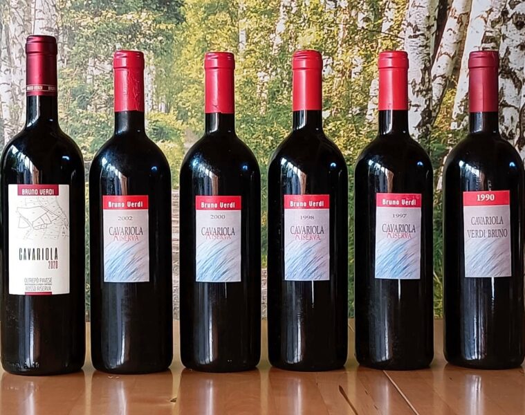 Six dark wine bottles with red foilcaps lined up on a wooden surface; labels read Bruno Verdi Cavariola Riserva with years 1990s–2002, set against a forest backdrop.
