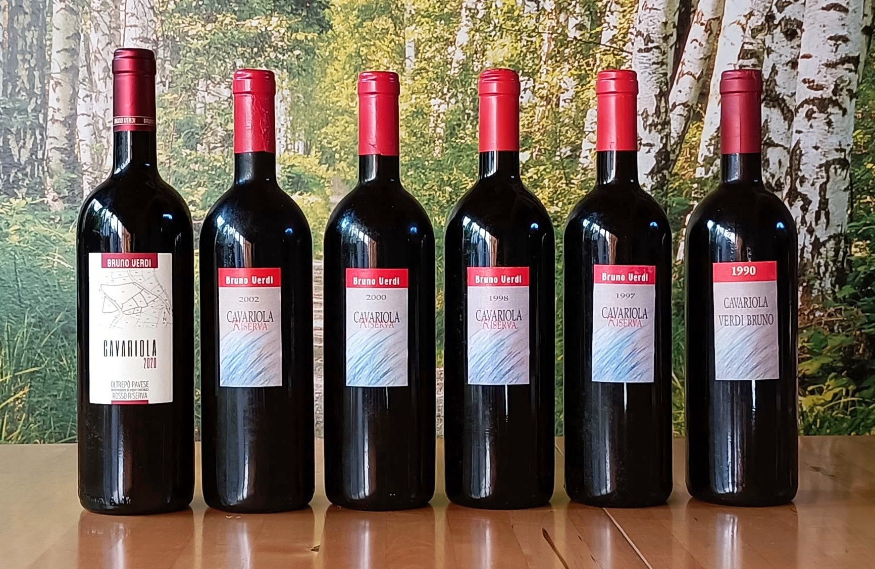 Six dark wine bottles with red foilcaps lined up on a wooden surface; labels read Bruno Verdi Cavariola Riserva with years 1990s–2002, set against a forest backdrop.