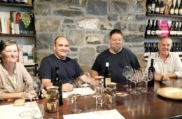 Four adults sit at a wine-tasting table with bottles, glasses, and a stone wall backdrop, smiling at the camera.