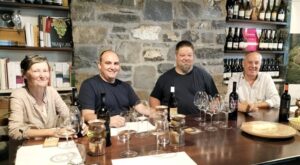 Four adults sit at a wine-tasting table with bottles, glasses, and a stone wall backdrop, smiling at the camera.