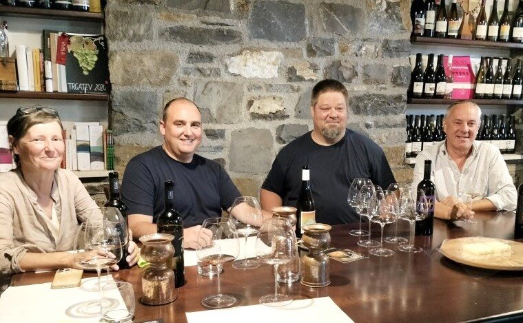 Four adults sit at a wine-tasting table with bottles, glasses, and a stone wall backdrop, smiling at the camera.
