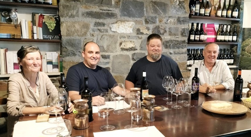 Four adults sit at a wine-tasting table with bottles, glasses, and a stone wall backdrop, smiling at the camera.