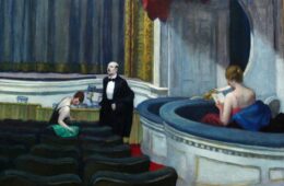 Theater stage scene: a man in a tuxedo stands center on stage while a woman in green sits among empty seats and another woman in a red dress reads on a curved balcony.