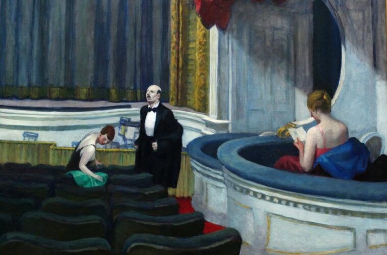 Theater stage scene: a man in a tuxedo stands center on stage while a woman in green sits among empty seats and another woman in a red dress reads on a curved balcony.