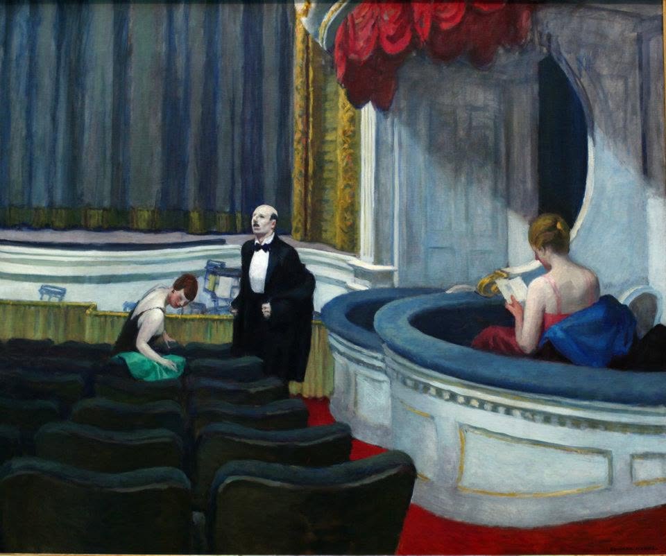 Theater stage scene: a man in a tuxedo stands center on stage while a woman in green sits among empty seats and another woman in a red dress reads on a curved balcony.