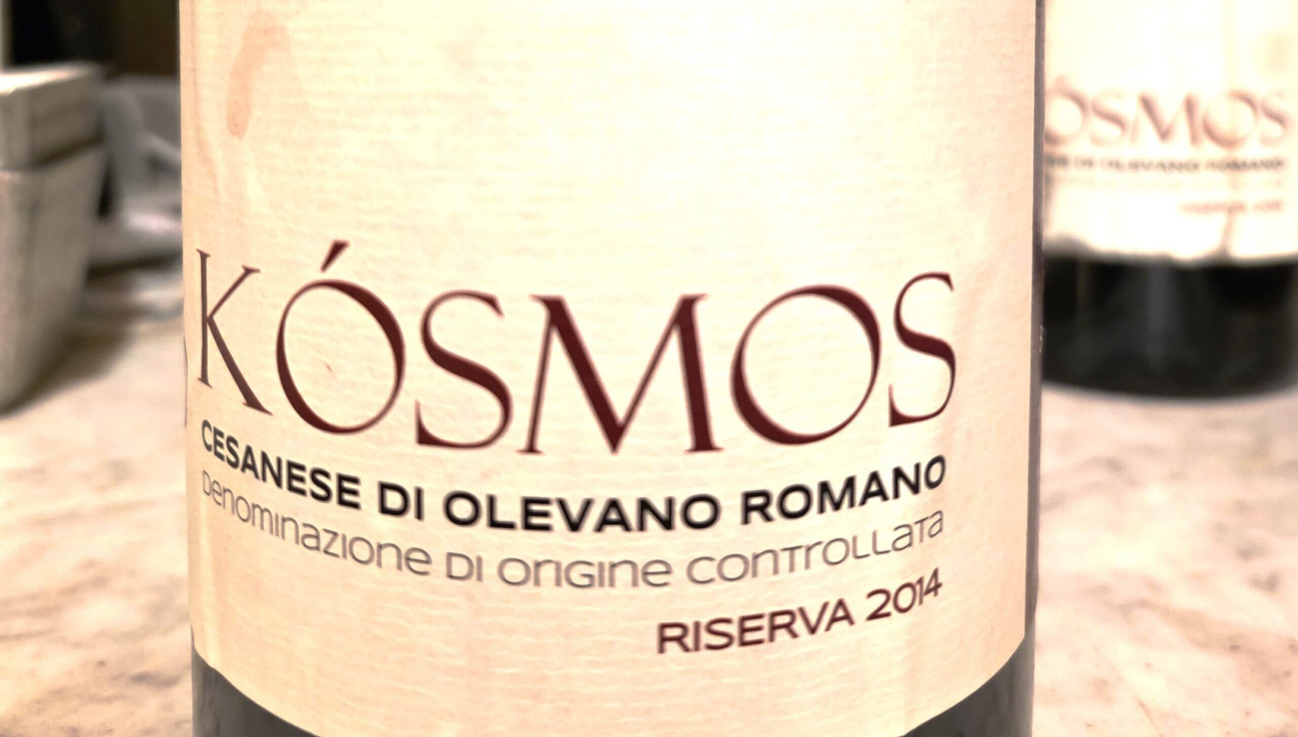 Close-up of a wine bottle label with the large word 'Kosmos' and smaller Italian origin text (Cesanese di Olevano Romano).