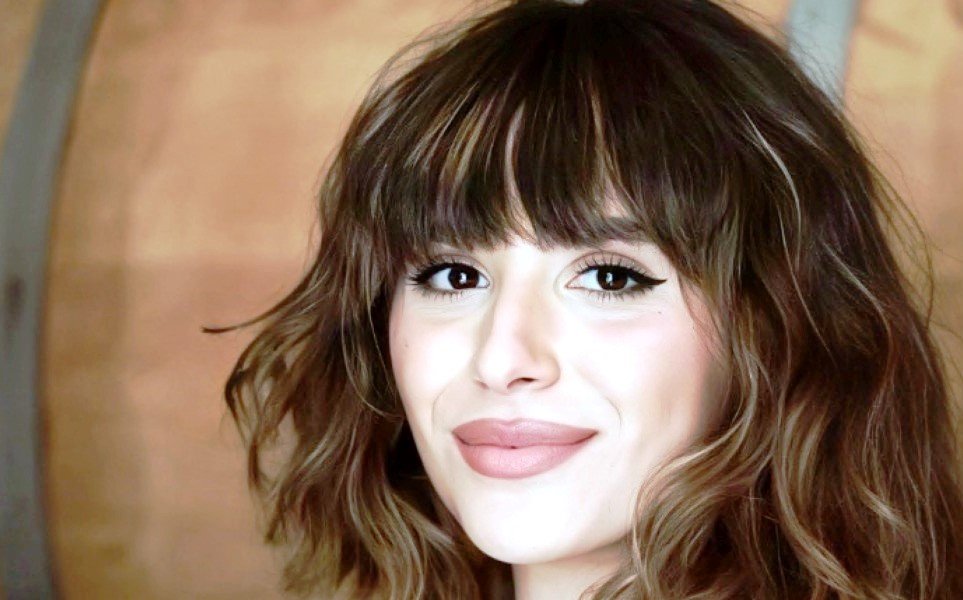 Close-up of a woman with wavy brown hair and bangs, smiling at the camera with defined eyeliner and pink lipstick.