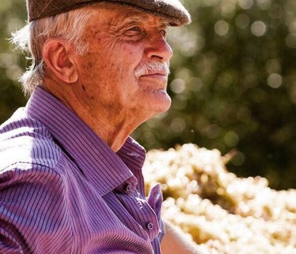 Older man in a brown flat cap and purple striped shirt, outdoors, looking to the right.