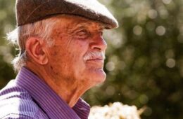 Older man in a brown flat cap and purple striped shirt, outdoors, looking to the right.