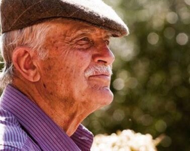 Older man in a brown flat cap and purple striped shirt, outdoors, looking to the right.