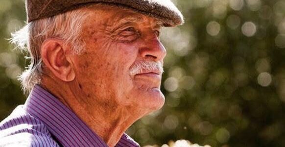 Older man in a brown flat cap and purple striped shirt, outdoors, looking to the right.