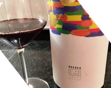 Glass of red wine beside a colorful patterned wine bottle with a limited edition label (Rockea) on a dark countertop.