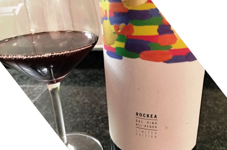 Glass of red wine beside a colorful patterned wine bottle with a limited edition label (Rockea) on a dark countertop.