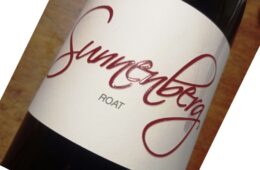 Close-up of a diagonal wine bottle with a white label featuring bold red cursive text and small gray text beneath.