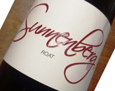 Close-up of a diagonal wine bottle with a white label featuring bold red cursive text and small gray text beneath.