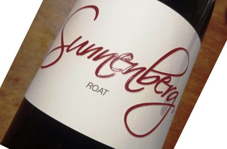 Close-up of a diagonal wine bottle with a white label featuring bold red cursive text and small gray text beneath.