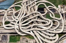 Aerial view of a sprawling concrete highway interchange with tangled looping ramps forming a maze-like pattern over roads and a river nearby