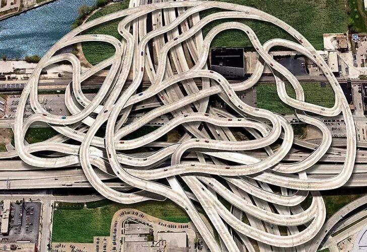 Aerial view of a sprawling concrete highway interchange with tangled looping ramps forming a maze-like pattern over roads and a river nearby