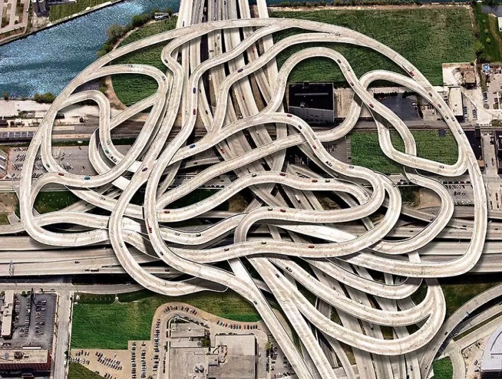 Aerial view of a sprawling concrete highway interchange with tangled looping ramps forming a maze-like pattern over roads and a river nearby