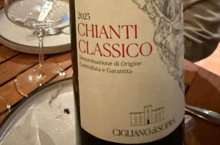Close-up of a 2023 Chianti Classico wine bottle label with a hillside village illustration on the label, on a dining table with a glass nearby.