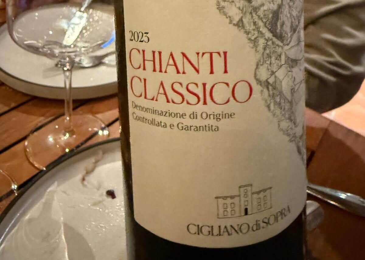 Close-up of a 2023 Chianti Classico wine bottle label with a hillside village illustration on the label, on a dining table with a glass nearby.