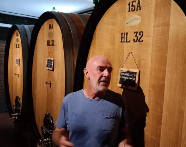 Man in a blue t-shirt stands beside large wooden wine barrels in a cellar, with small chalkboard signs hanging on the barrels.