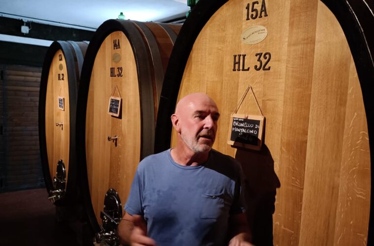 Man in a blue t-shirt stands beside large wooden wine barrels in a cellar, with small chalkboard signs hanging on the barrels.
