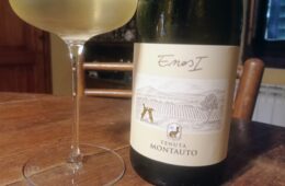 Wine glass beside a pale white wine bottle on a wooden table, label showing Tenuta Montauto.