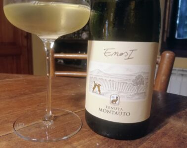 Wine glass beside a pale white wine bottle on a wooden table, label showing Tenuta Montauto.