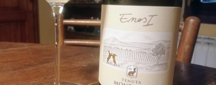 Wine glass beside a pale white wine bottle on a wooden table, label showing Tenuta Montauto.