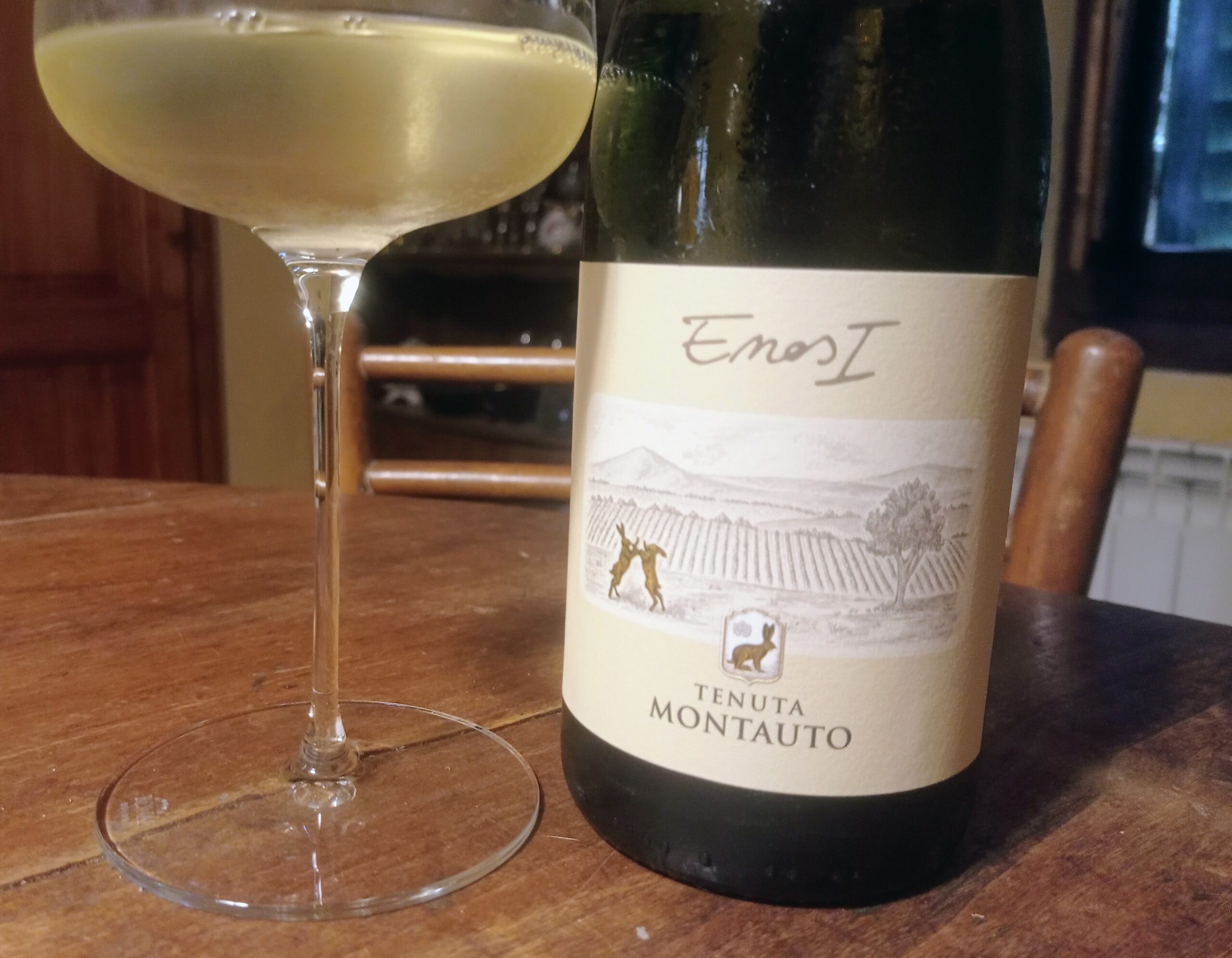 Wine glass beside a pale white wine bottle on a wooden table, label showing Tenuta Montauto.