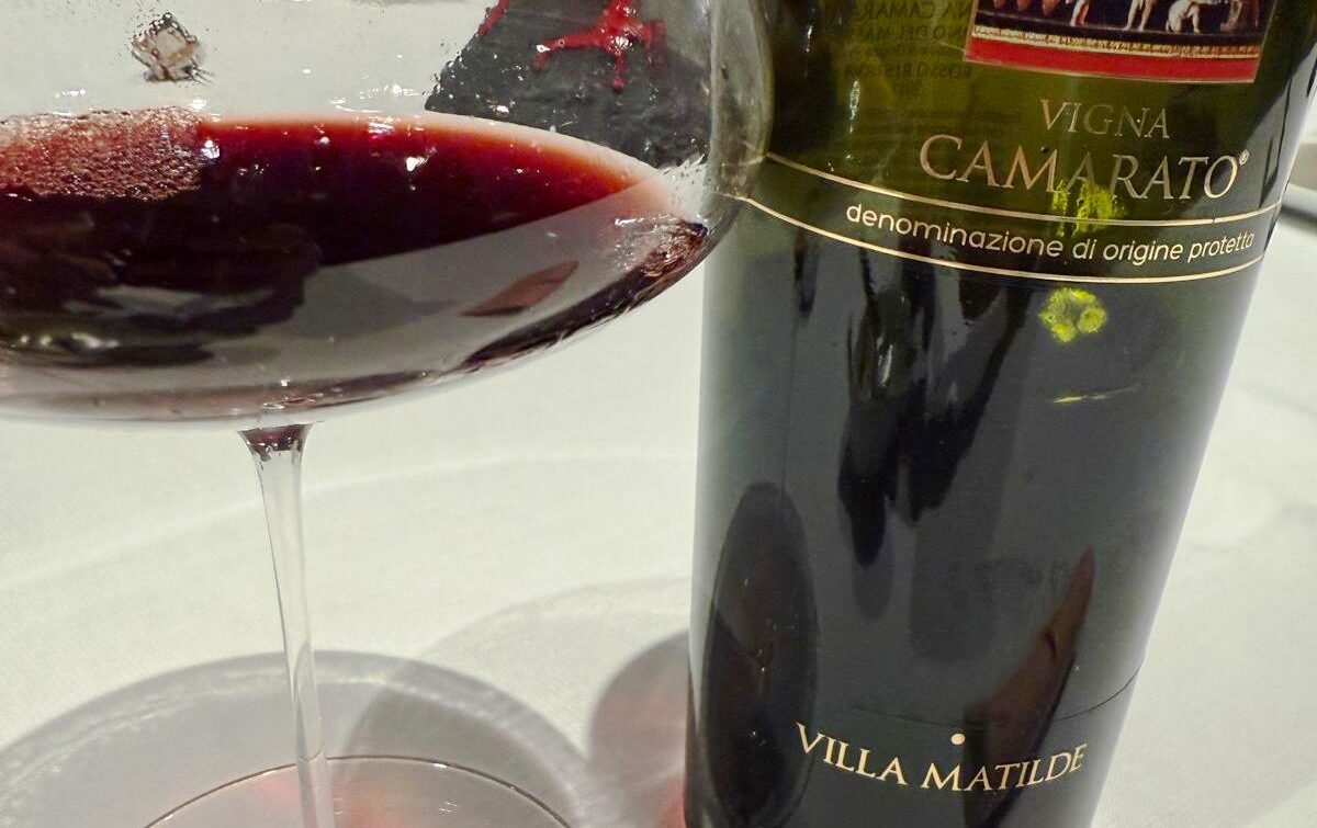 Wine bottle labeled Villa Matilde, Vigna Camarato, gently poured red wine in a glass beside it on a white table.