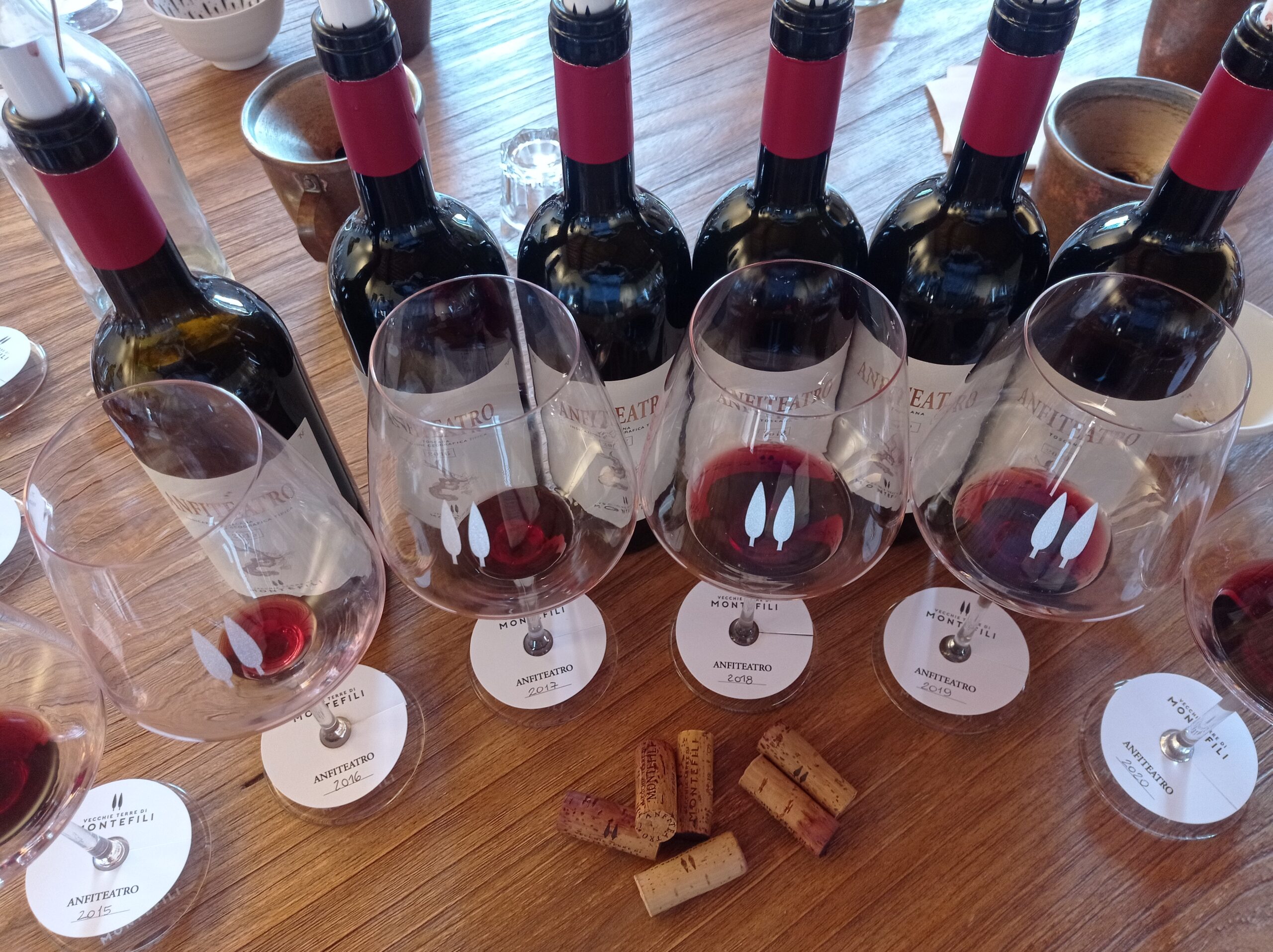 Row of wine bottles with red capsules and glasses on a wooden table, labels reading ANFITEATRO and corks nearby—likely a tasting setup.