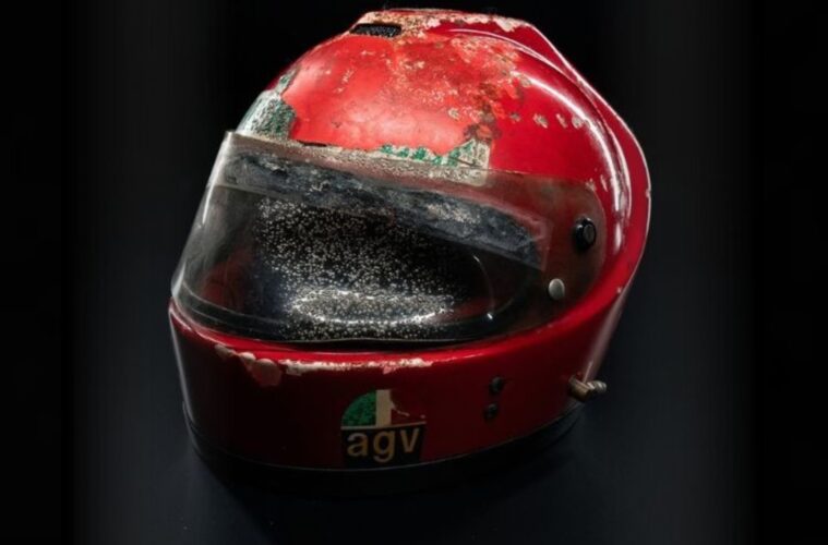 Close-up of a red racing helmet with a dirty, scratched visor against a dark background.