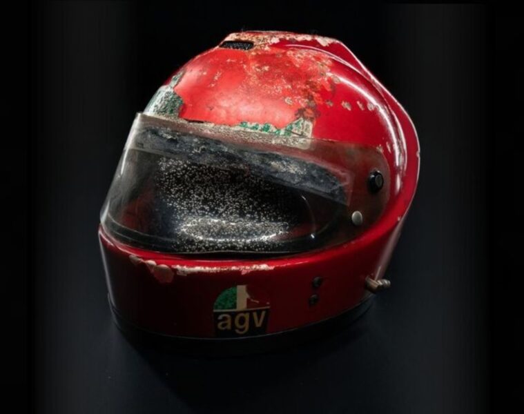 Close-up of a red racing helmet with a dirty, scratched visor against a dark background.