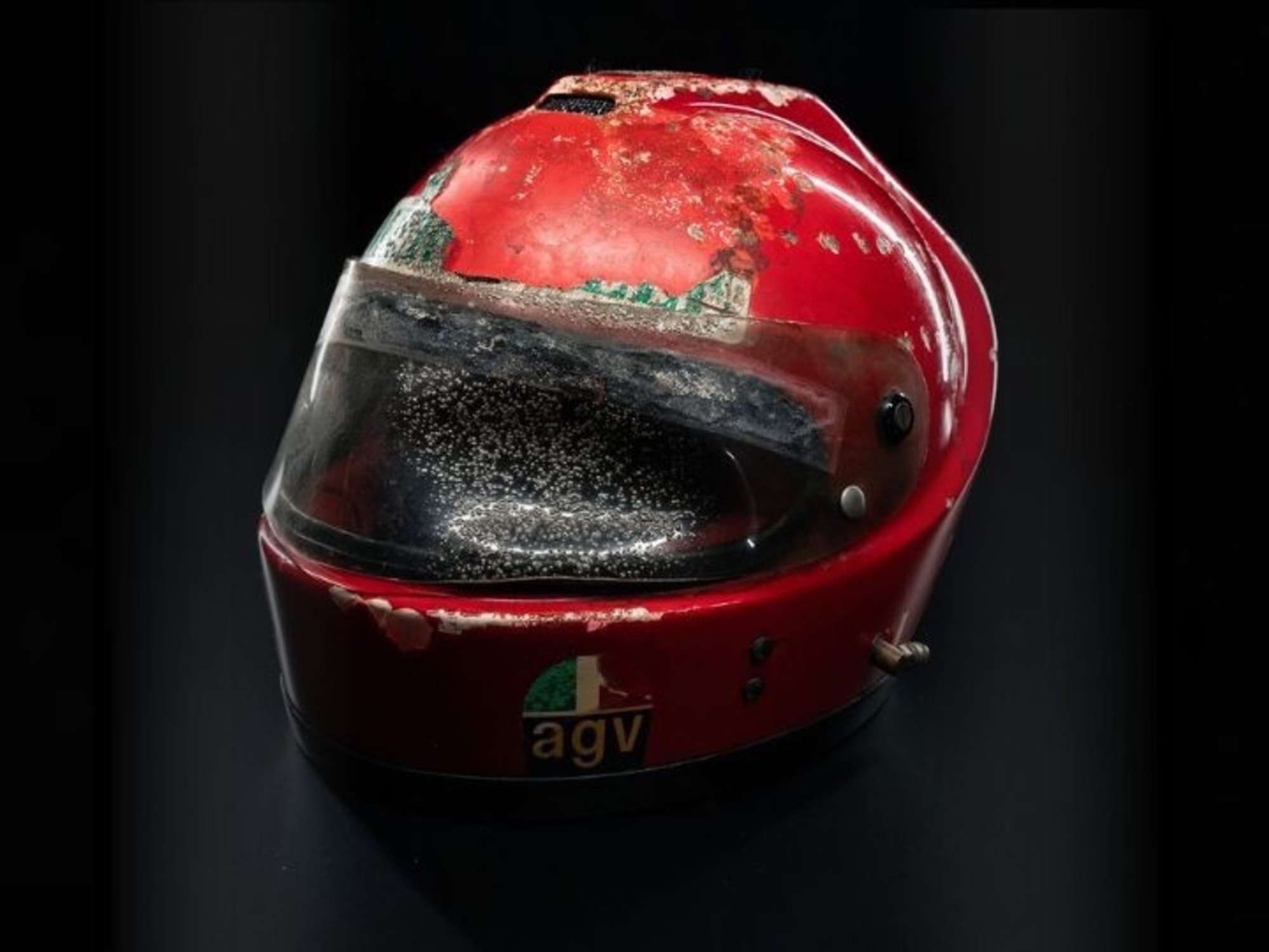 Close-up of a red racing helmet with a dirty, scratched visor against a dark background.