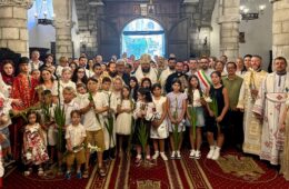 Group photo inside a stone church with children and clergy holding white lilies during a ceremony.