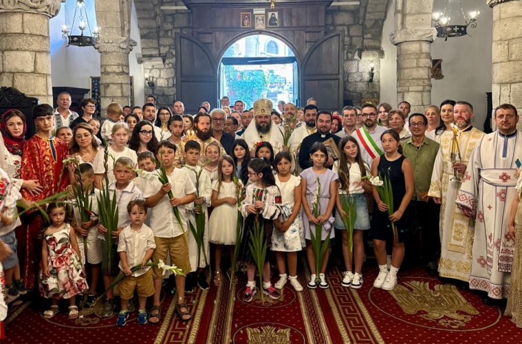 Group photo inside a stone church with children and clergy holding white lilies during a ceremony.
