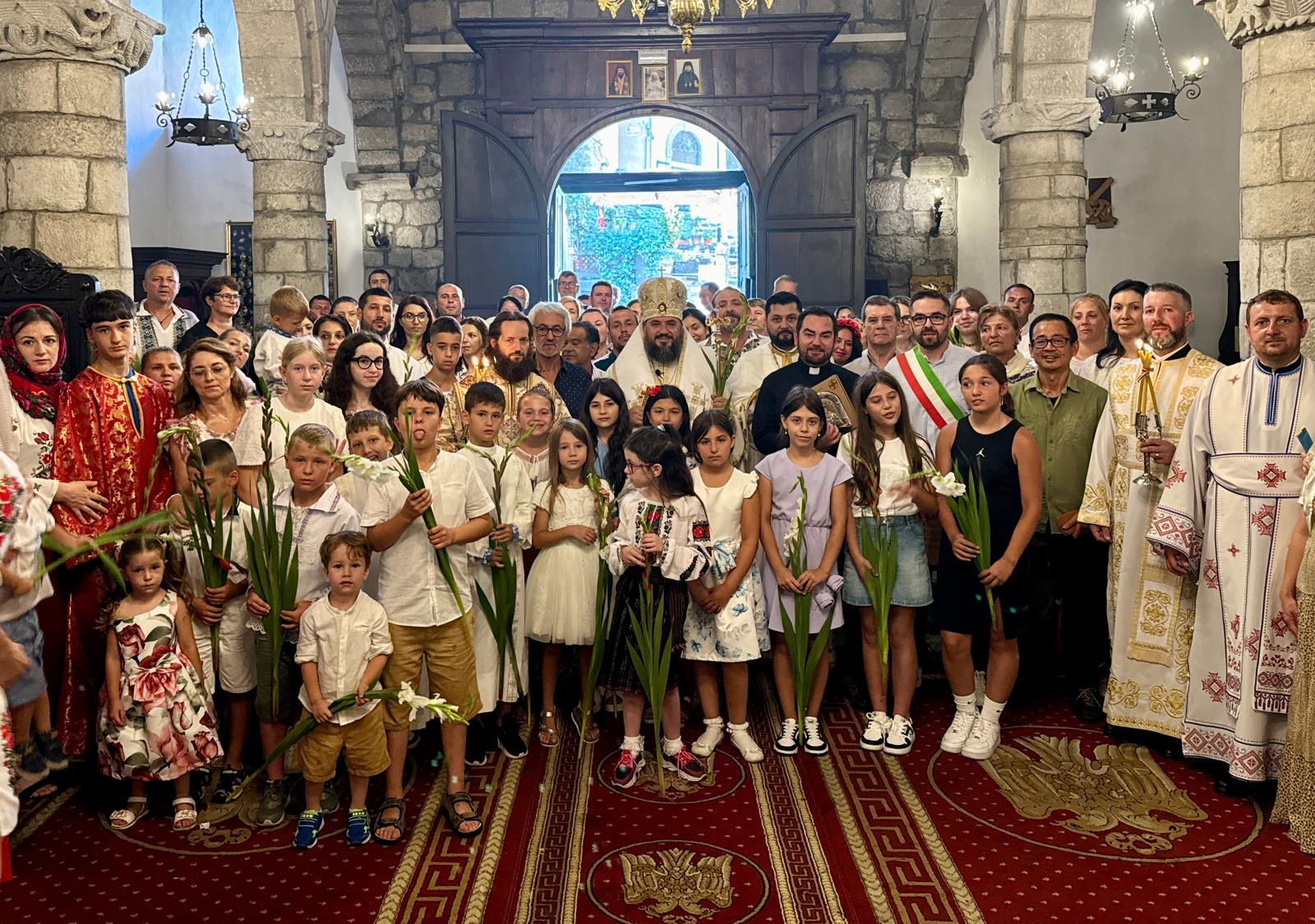 Group photo inside a stone church with children and clergy holding white lilies during a ceremony.